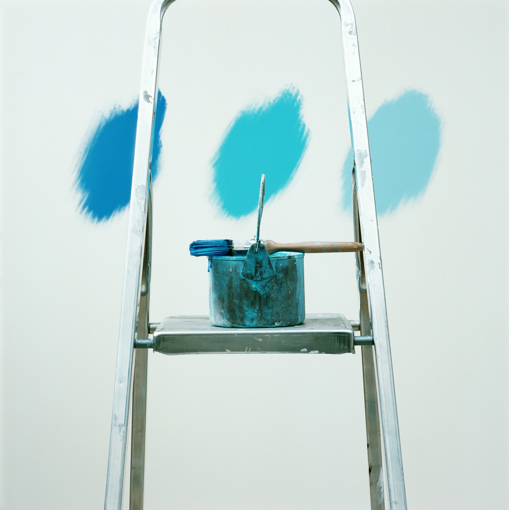 Balde de tinta e pincel, molhado com tinta azul, sobre uma escada. Na parede, 3 tons de azul.