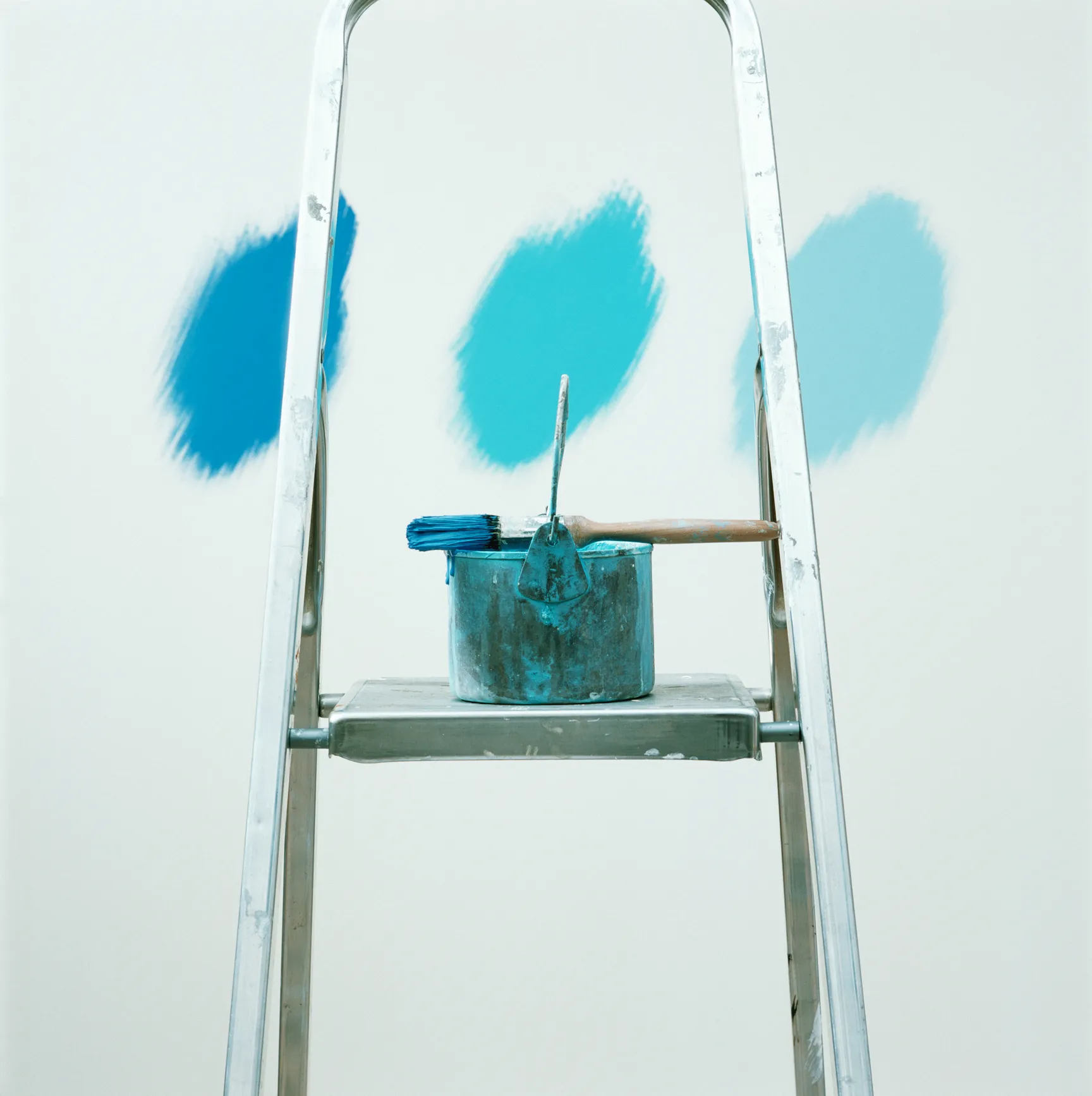 Balde de tinta e pincel, molhado com tinta azul, sobre uma escada. Na parede, 3 tons de azul.