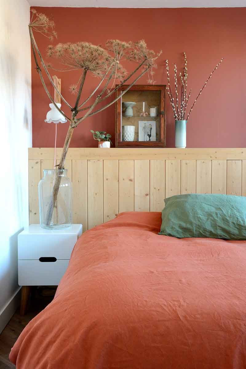 Quarto de casal com tons terrosos. A cama possui colcha vermelha e cabeceira de madeira pinus crua.