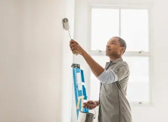 Homem pintando uma parede com rolo. Atrás dele estão uma escada e uma janela recebendo a luz do dia.