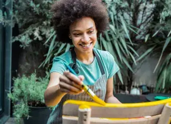 Mulher sorrindo, usando um avental e pintando uma cadeira de amarelo.