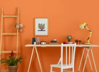 Parede laranja com um quadro e, na frente, uma mesa para estudos contendo diversos itens.