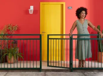 Mulher sorrindo e com as mãos sobre o portão. Atrás está sua casa, com a parede vermelha.